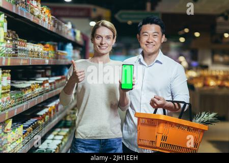 Ritratto di una coppia asiatica felice di consumatori, acquirenti in un supermercato o negozio di alimentari, guardando la macchina fotografica, sorridendo. Stanno tenendo un telefono e mostrando uno schermo verde chromakey Foto Stock