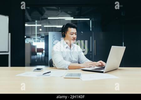 Giovane uomo d'affari asiatico che lavora su un portatile in cuffie con microfono. Seduta su una scrivania in un ufficio moderno. Serio, concentrato. Foto Stock