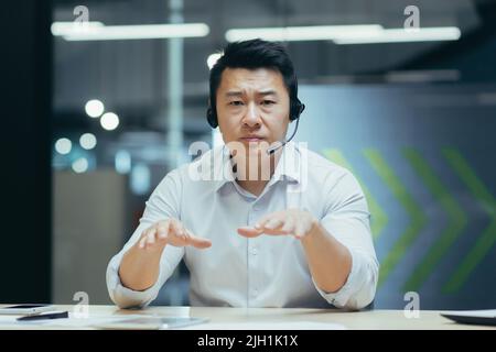 Pullman per affari. Un giovane uomo asiatico in cuffie con microfono conduce l'allenamento professionale con la fotocamera, spiega, dice, sorride. Seduto dietro una scrivania in un ufficio moderno. Foto Stock