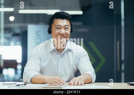 Pullman per affari. Un giovane uomo asiatico in cuffie con microfono conduce l'allenamento professionale con la fotocamera, spiega, dice, sorride. Seduto dietro una scrivania in un ufficio moderno. Foto Stock