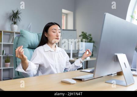 Pausa e meditazione al lavoro. Giovane donna d'affari asiatica bella che indossa occhiali seduti alla scrivania in ufficio con gli occhi chiusi, tenendo le mani in posa di lotus yoga, riposo, rilassato. Foto Stock