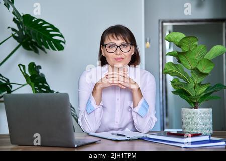 Donna di mezza età che guarda la macchina fotografica seduta alla sua scrivania con il laptop Foto Stock