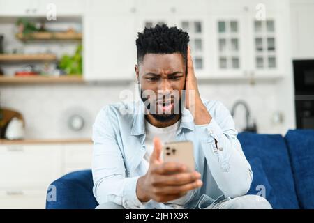 Arrabbiato ha stressato uomo multirazziale che ha sgradevole conversazione di videochiamata, parlare su smartphone online e discutere, mentre si siede sul divano e guardando lo schermo del telefono con espressione del volto frustrata Foto Stock
