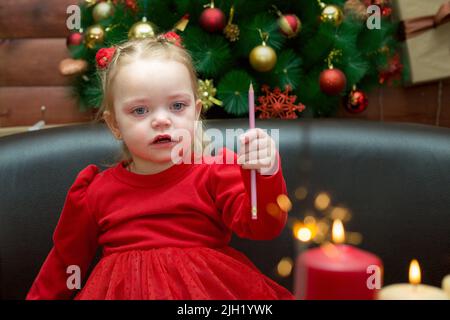 Una ragazza tristemente tristemente, di tre anni in un vestito rosso con una matita in mano sullo sfondo di un albero di Natale teso. Foto Stock