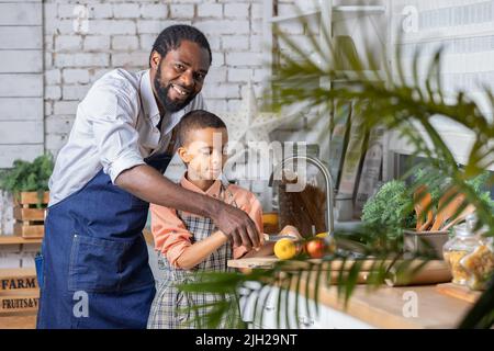 Il padre nero e il bambino che cucinano verdure fresche in cucina a casa. Papà africano e bambino ragazzo che si preparano insieme. Foto Stock