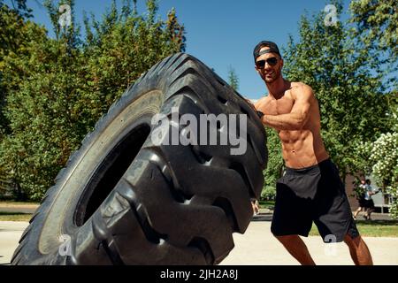 Allenamento uomo forte sollevamento grande palestra all'aperto pneumatico. Sportivo giovane adulto caucasico ragazzo flip ruota grande sulla natura. Attrezzature sportive fatte a mano Foto Stock