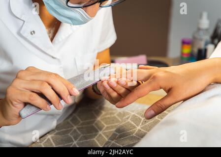 Manicurista con misure di sicurezza e maschera facciale che lavora sulle unghie del cliente al manicure e al salone pedicure dopo la riapertura della pandemia Foto Stock