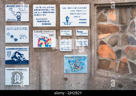nagasaki, kyushu - dicembre 11 2021: Desideri di pace e illustrazioni su placche di ceramica sul muro di un monumento a forma di fiamma olimpica greca di p. Foto Stock
