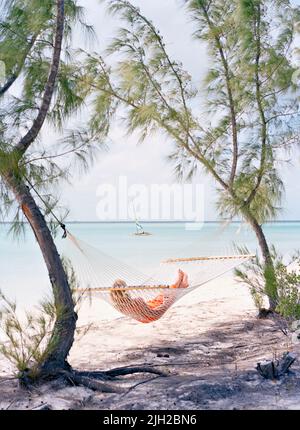 Un ospite si rilassa in un'amaca sulla spiaggia di Tiamo mentre un catamarano naviga vicino. Isola di Andros del sud, Bahamas Foto Stock