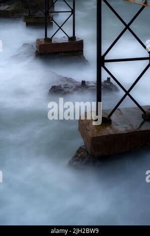 Il surf atlantico sfugge a una nebbiolina fine mentre si aggira avanti e indietro sotto la lunga passerella di ferro che conduce al Rocher de la Vierge o Rocca della Vergine a Biarritz, Nouvelle-Aquitaine, Francia. Il ponte sulla baia di Biscaglia è stato progettato nel 1887 dall'ingegnere civile francese Gustave Eiffel. Foto Stock