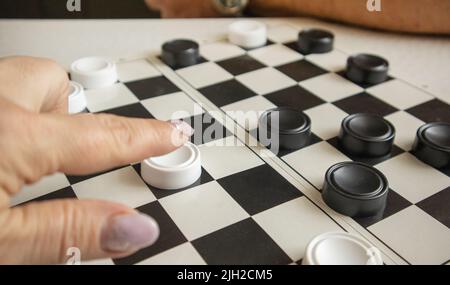 La mano di una donna muove una pedina bianca su un campo da gioco in bianco e nero, il concetto di hobby e giochi per la casa. Foto Stock