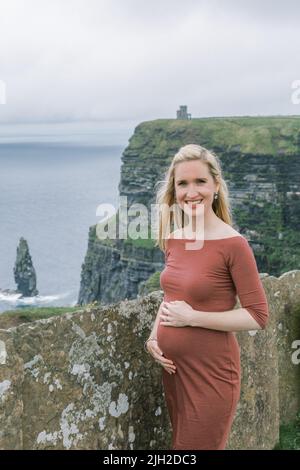Donna incinta in abito rosso si erge alle scogliere di Moher in Irlanda Foto Stock