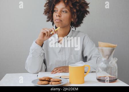 La donna afro-americana?urly sta facendo colazione e sta scrivendo un diario. Foto Stock