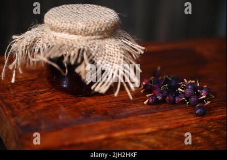 Primo piano di un vasetto di marmellata fatta in casa di uva spina e frutti di bosco freschi maturi su una superficie rustica in legno. Concetto di inscatolamento Foto Stock