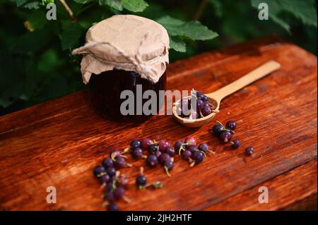 Ancora vita di un vaso di un confit fatto in casa, cucchiaio di legno e frutti di bosco su una superficie rustica di legno. Tradizioni culinarie Foto Stock