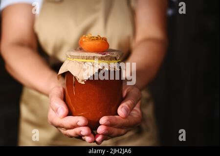 Dettagli: Una casalinga che tiene un vaso di marmellata di albicocche fatta in casa. Un succoso frutto maturo di albicocca sul coperchio Foto Stock