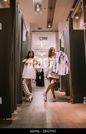 Due belle ragazze in piedi alla stanza di cambiamento nel negozio che provano i vestiti Foto Stock