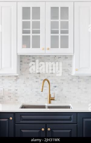 Un dettaglio lavandino della cucina girato in una cucina bianca e blu con un rubinetto d'oro, piano di appoggio in marmo, e un piccolo backsplash di piastrelle di marmo della metropolitana. Foto Stock