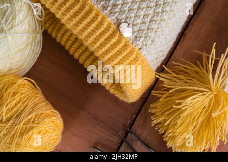 cappellino giallo e bianco per l'inverno, lavorato a mano a crochet su fondo di legno arancione, accanto ad esso due rotoli di lana e tre aghi di acciaio pronti Foto Stock