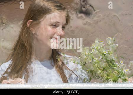 Bella ragazza che guarda attraverso la finestra con gocce di pioggia sul vetro con un bouquet di fiori camomilla, primo piano. Felice ragazza giovane guarda fuori finestra Foto Stock