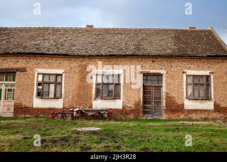 Foto di una fattoria abbandonata in Vojvodina, in Serbia, con la facciata della sua casa principale gravemente danneggiata e decadente. Foto Stock