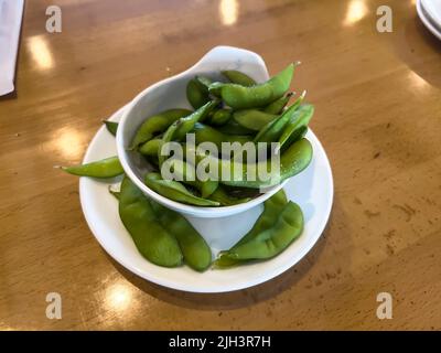 Primo piano, focalizzazione selettiva su cialde di edamame in una piccola tazza bianca su un tavolo di legno all'interno di un ristorante Foto Stock