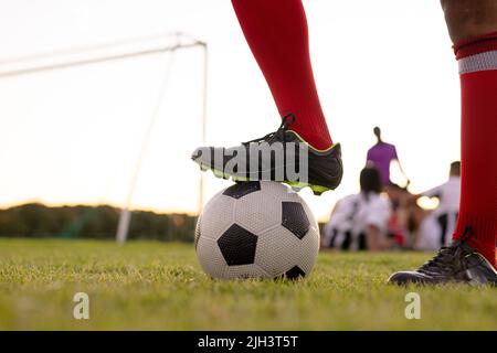 Bassa sezione di atleta caucasico maschio indossando calze rosse e scarpe nere mettendo gamba su pallone da calcio Foto Stock