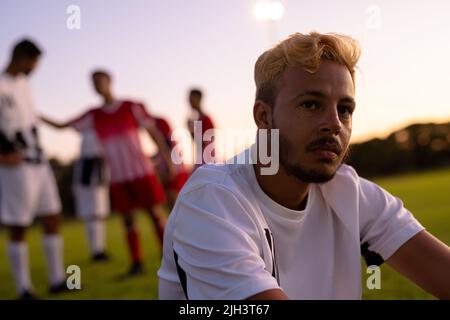 Caucasico giocatore di calcio maschile premuroso guardando lontano e seduto contro il cielo limpido al parco giochi Foto Stock