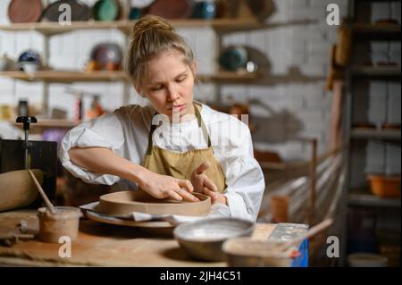 Un ceramista fa un piatto. Donna in grembiule lavora in un laboratorio di ceramica. Foto Stock