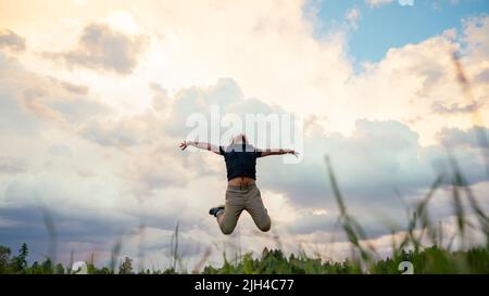 Vista ad angolo basso attraverso le lame di erba di un giovane gioioso che salta in alto sotto un cielo serale. Foto Stock