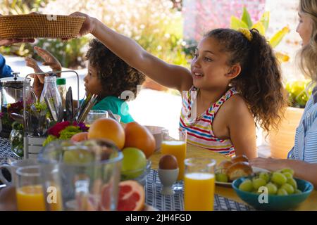 Ragazza carina multirazziale passando cibo mentre fare colazione con madre e fratello al tavolo da pranzo Foto Stock