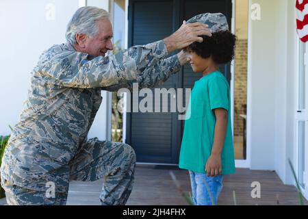 Nonno militare caucasico in abbigliamento camouflage con cappuccio al nipote biraciale fuori casa Foto Stock