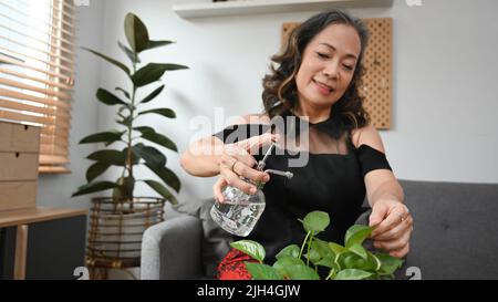 Donna sorridente matura innaffiare piante domestiche in soggiorno. Concetto di cura di piante e lavori domestici Foto Stock