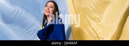 giovane donna ucraina allegra in colore block vestire posando vicino bandiera blu e gialla, bandiera Foto Stock