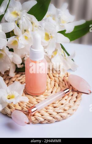 Bottiglia di siero rosa e rullo facciale in quarzo rosa con grandi fiori bianchi. Bellezza, spa e concetto di benessere. Primo piano. Foto Stock