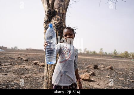 Bella bambina africana in piedi in un campo di pietra asciutta che tiene su una bottiglia vuota di plastica che simboleggia la scarsità d'acqua nella regione subsahariana Foto Stock