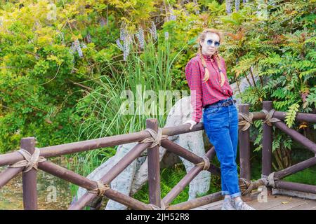 Una donna in una camicia a quadri e jeans si erge su una ringhiera di legno vicino ad un laghetto in un parco di primavera tra piante verdi. Fine settimana a piedi. Moda giovanile. Foto Stock