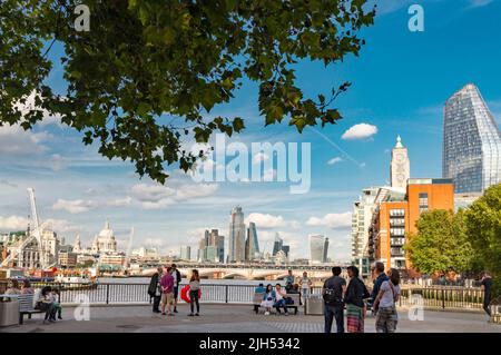 Londra,Inghilterra,UK-Luglio 21 2019: I giovani si radunano all'ombra degli alberi, lungo la passeggiata lungo il fiume, ammirando l'iconico skyline di Londra attraverso il Foto Stock