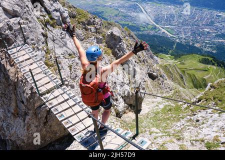 Ragazza su Nordkette via ferrata ponte di legno solleva le braccia, guarda verso il basso sopra Innsbruck città, Austria. Klettersteig, casco, avventura, attivo, estate. Foto Stock