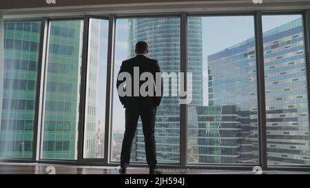 Un uomo d'affari in un vestito si erge alla finestra sullo sfondo dei grattacieli. Un uomo d'affari riuscito in un vestito si leva contro lo sfondo di Foto Stock