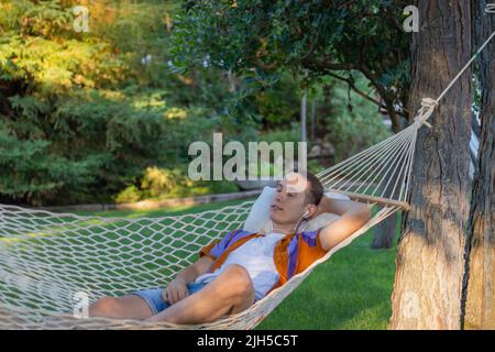 Giovane uomo in vacanza sdraiato in un'amaca mentre ascolti musica tranquilla e rilassante con la natura in background in estate Foto Stock
