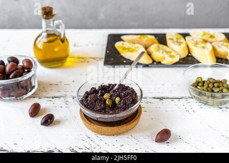 Piatto di olive in ciotola e ingredienti di base - olio d'oliva, olive e capperi su sfondo bianco di legno Foto Stock