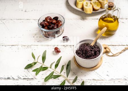 Piatto di olive in ramequina e ingredienti di base - olio d'oliva, olive su sfondo bianco di legno con crostini e rametti d'oliva Foto Stock