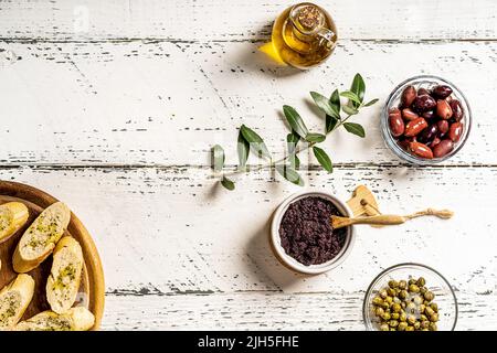 Patata di olive con ingredienti - olio d'oliva, olive, capperi su sfondo bianco di legno con rametti d'oliva e crostini Foto Stock