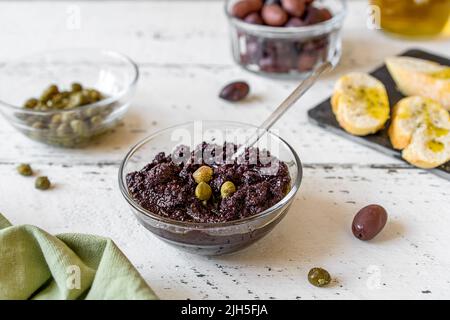 Piatto di olive in ciotola e ingredienti di base - olio d'oliva, olive e capperi su sfondo bianco di legno Foto Stock