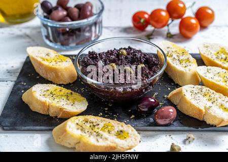 Piatto di olive in ciotola con crostini su tagliere di pietra nera e ingredienti di base a parte - olive, olio, capperi. Foto Stock