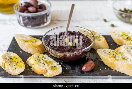 Piatto di olive in ciotola con crostini su tagliere di pietra nera e ingredienti di base a parte - olive, olio, capperi. Foto Stock