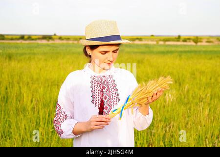 Defocus giovane donna in vyshyvanka e cappello che tiene bouquet di pikelets d'oro maturo di grano legato sul prato sfondo natura. Bandiera Ucraina Foto Stock