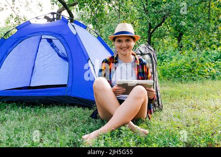 Defocus giovane donna che lavora sul tablet vicino campeggio tenda all'aperto circondato da bella natura. Freelance, sabbatico, salute mentale. Fuori fuoco. Foto Stock