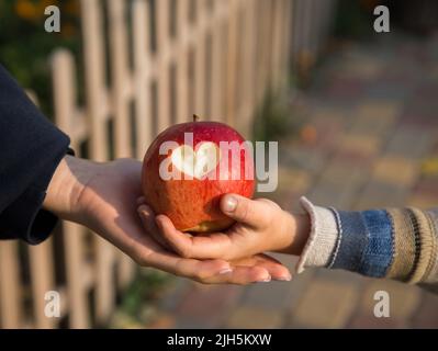 la mano femminile e la mano di un bambino tengono una mela rossa grande con un cuore scolpito su di essa. Il concetto di relazioni amichevoli nella famiglia, nella madre, nella forma Foto Stock